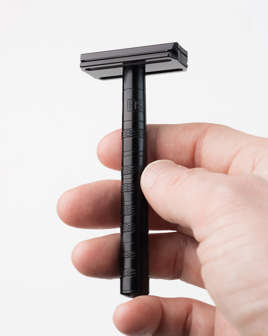 Hand holding a black Henson AL13 double-edge safety razor with knurled handle and HS logo, upright against a white background
