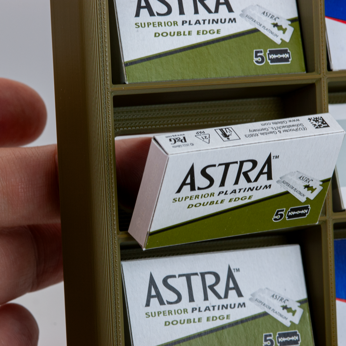 Army green 3D-printed razor blade display stand holding stacked boxes of Astra Superior Platinum double edge blades, with a hand pulling one box out