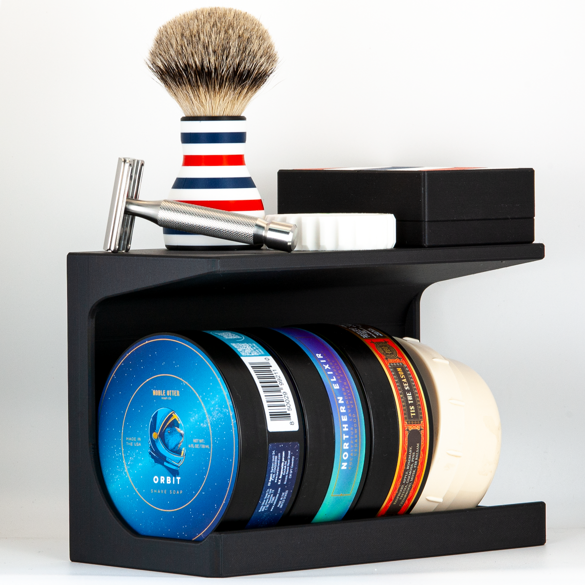Noble Otter shaving soaps displayed on a black soap stand with a razor, brush, storage case, and razor refueler on top