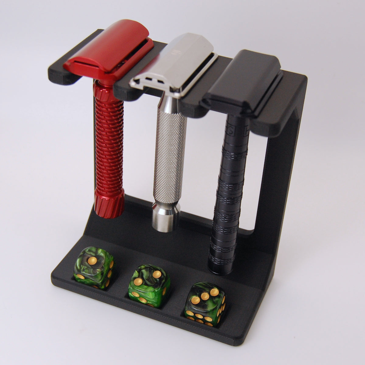 Black display stand with three safety razors in red, silver, and black, and three green marbled dice with gold pips in base