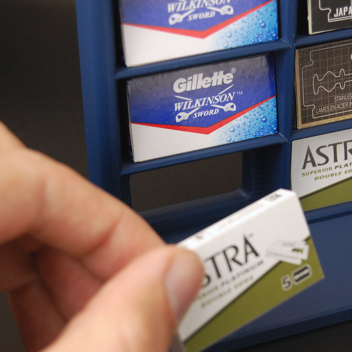 Navy blue razor blade display stand holding various blade tucks on black background, with a hand removing a tuck from its pocket slot.