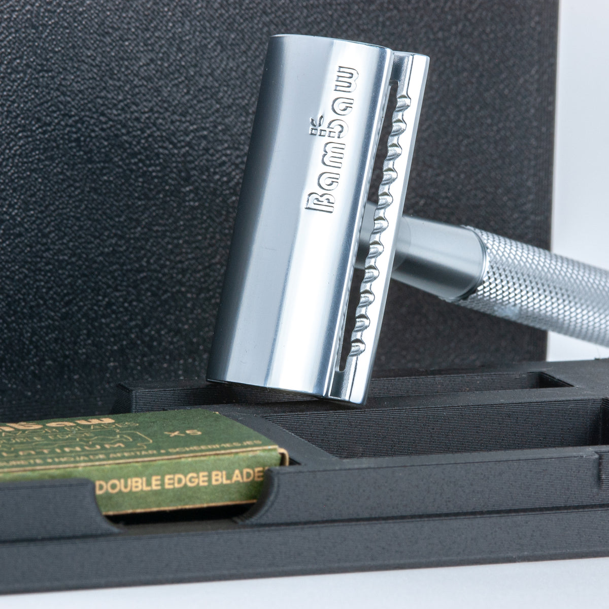Close-up of the chrome Bambaw safety razor head engraved with the Bambaw logo, in front of its travel case