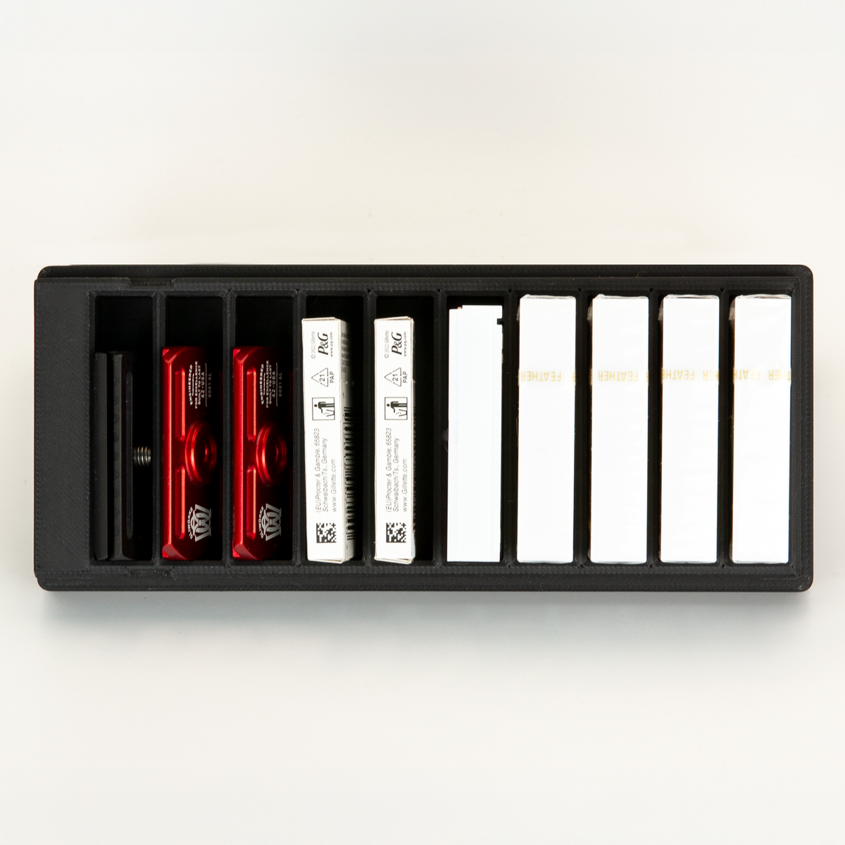 Black 10-slot razor blade box from above: a spare Blackland Blackbird head and two red razor plates in the first slots, seven blade boxes in the rest