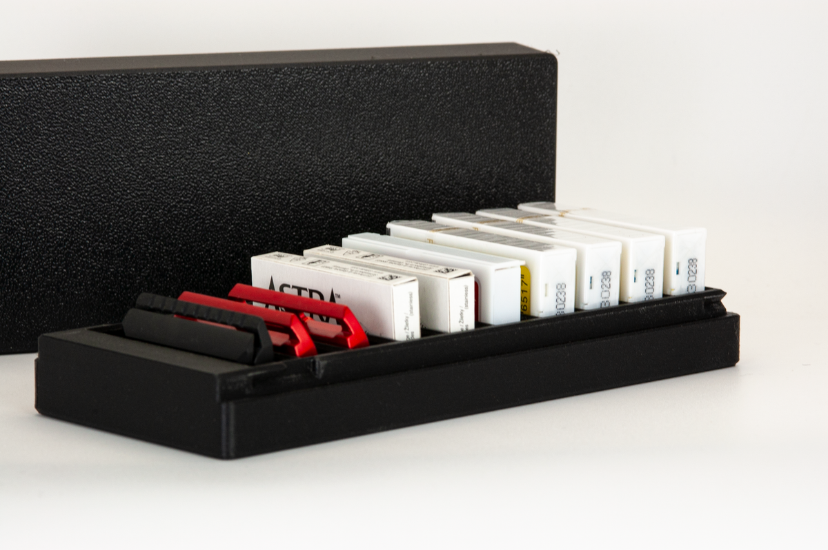 Open black razor blade box at an angle, lid behind: a spare Blackland Blackbird head and red razor plates in the first slots, Astra and Feather blade boxes in the rest