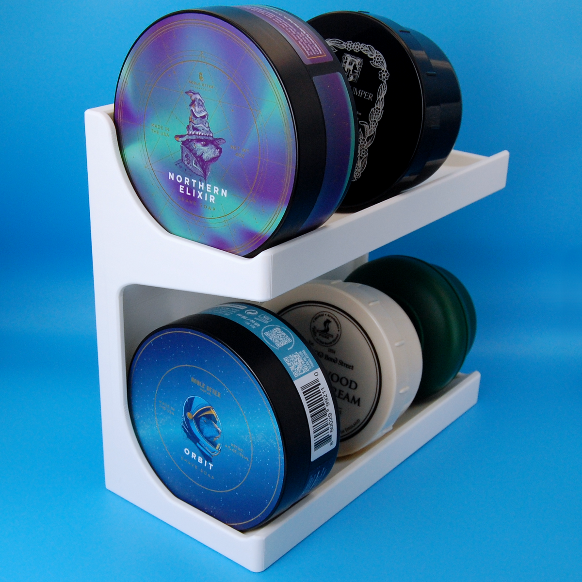 White shaving soap stand holding several shaving soaps, including Noble Otter Orbit and Northern Elixir, on a light blue background.